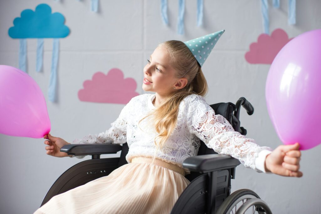 beautiful smiling girl in birthday cap sitting in wheelchair and holding balloons at birthday party